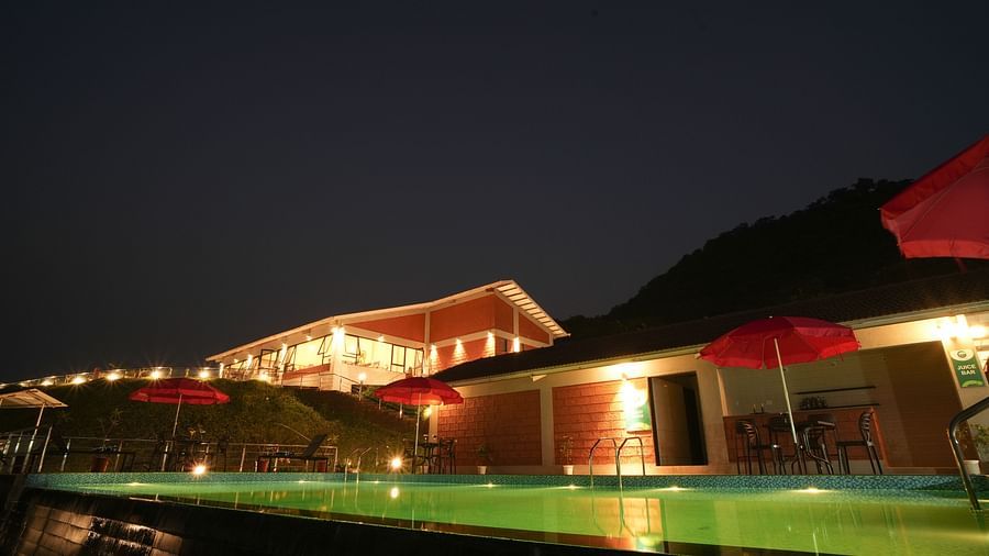 A swimming pool lit at night with red umbrellas and a building in the background - Gaurav Lords Resort Shrivardhan.