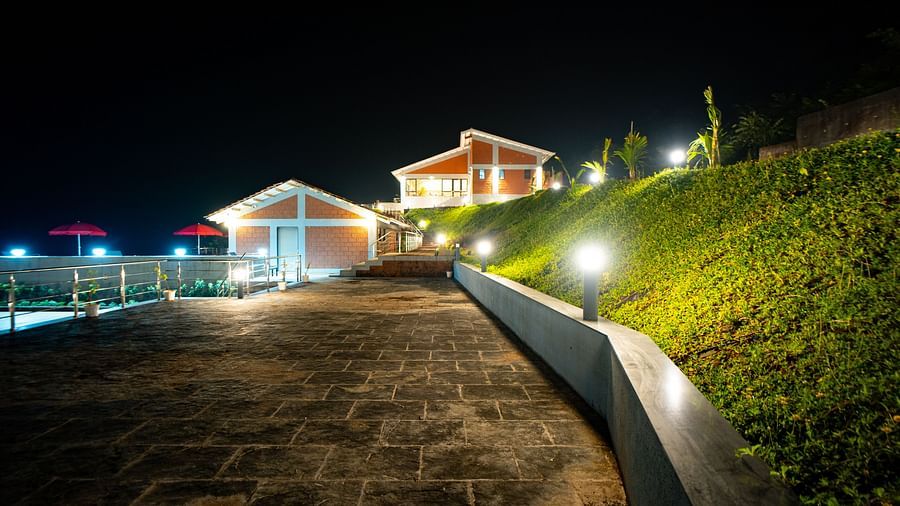 A stone pathway leading to illuminated buildings at night, with a green hillside on the right - Gaurav Lords Resort Shrivardhan.