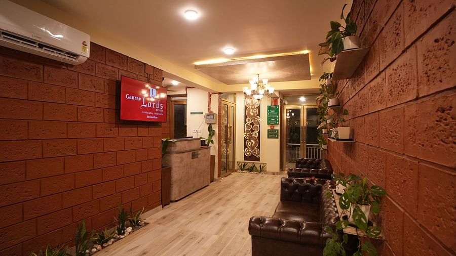 Interior hallway of a resort with textured red brick walls, a wooden reception desk, a dark leather sofa, and potted plants. A television is mounted on the wall - Gaurav Lords Resort Shrivardhan.