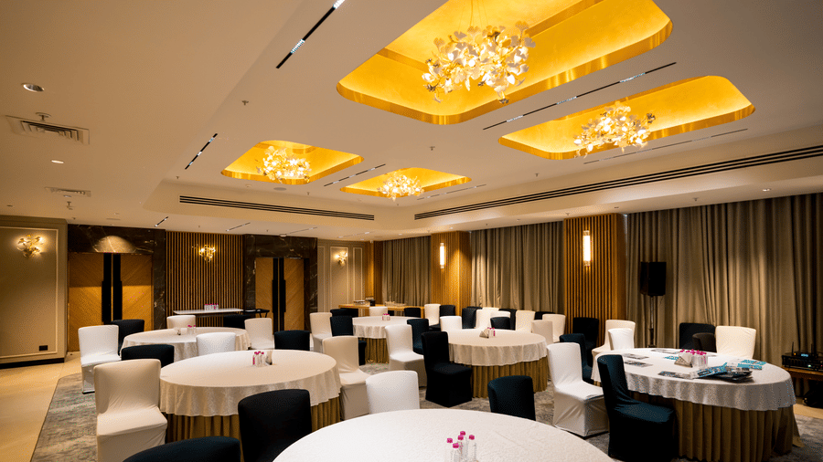 Banquet hall Galaxy 2 at Gemini Continental features round tables with tablecloths, black and white chairs, ceiling panels with lights, chandeliers, carpeted floor, curtains, wall lights, buffet counters, doors, speaker, cables, water bottles, and table it