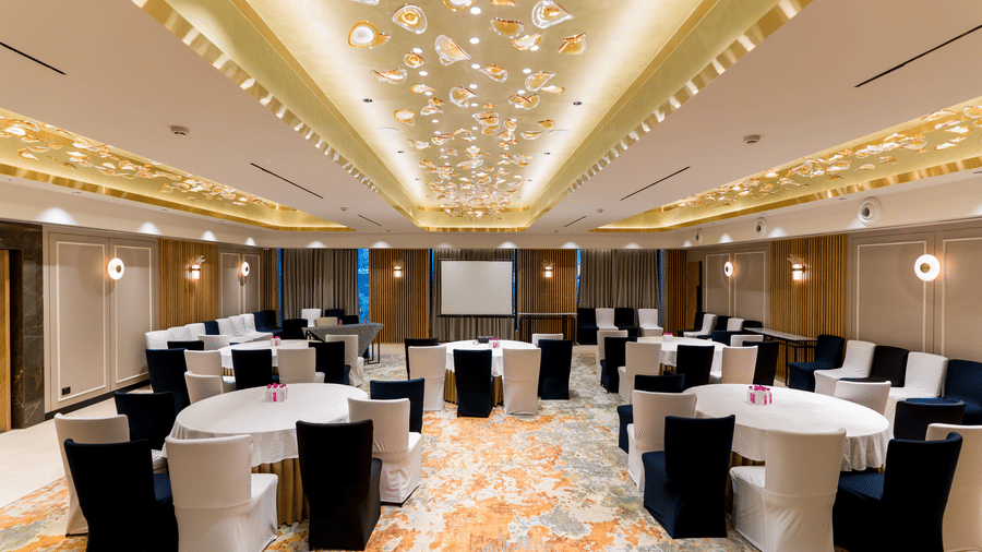 Conference hall Galaxy 1 at Gemini Continental featuring multiple round tables facing a front screen, accent walls, and a decorative ceiling. 