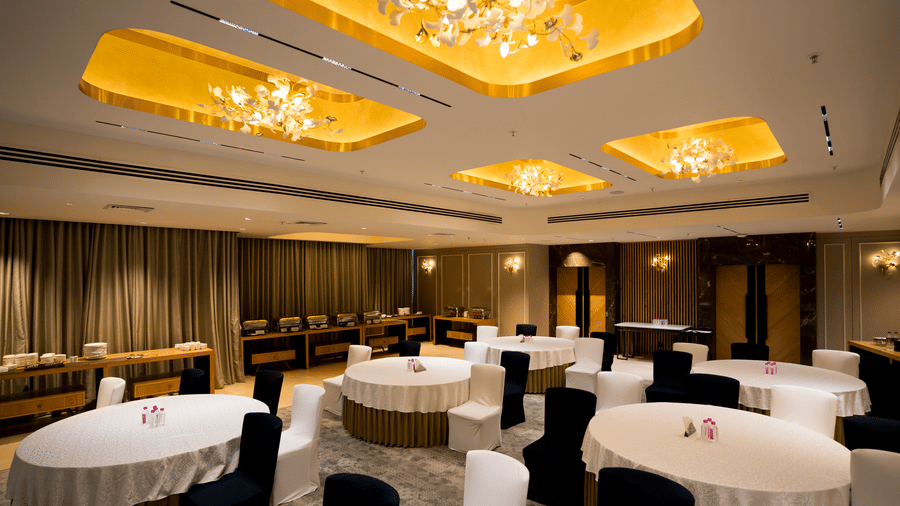 Banquet hall Galaxy 2 at Gemini Continental with round tables, tablecloths, black and white chairs, ceiling panels with lights, chandeliers, curtains, carpeted floor, wall lights, buffet counters, doors, and table items.