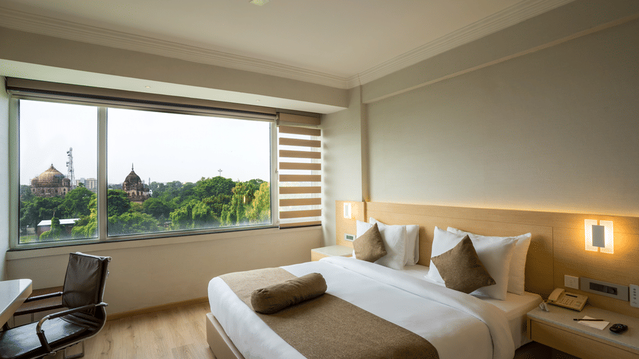 Bedroom of a Suite at Gemini Continental with a large window overlooking trees and historic buildings, featuring a desk and bedside lighting.