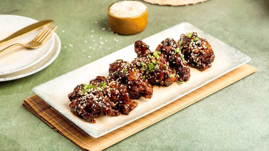 Glazed chicken wings topped with sesame seeds and green onions, served on a rectangular plate with tableware in the background, at Zaika in Gemini Continental.