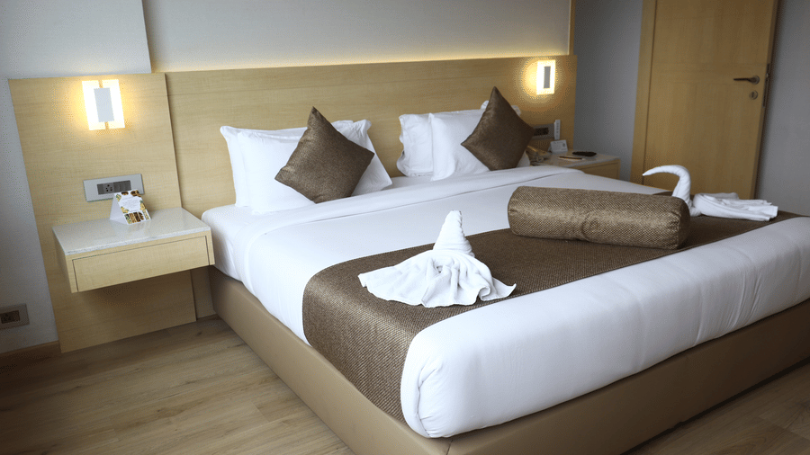 Suite bedroom with a bed dressed in white and brown linen, swan-shaped towel art, wall sconces on either side and light wooden flooring below.