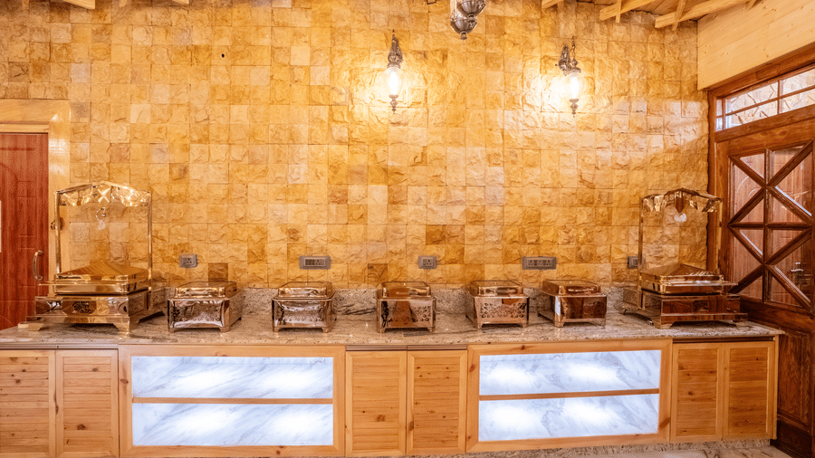 A modern buffet counter with stainless steel food warmers set against a golden stone-tiled wall - Golden Oak By The Lake Hill, Khurpatal
