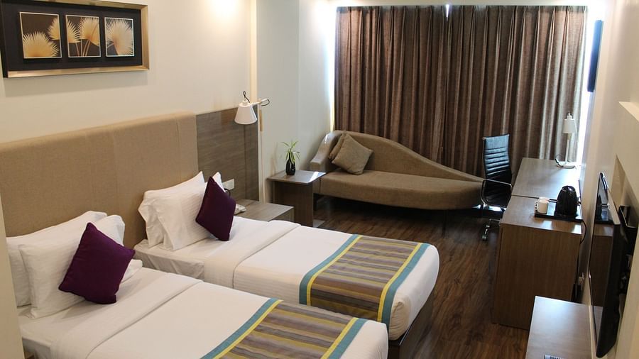 An image showing a pair of twin beds with lush decors, a long sofa with cushion, and a flat screen TV, inside a room with closed curtains.- Golden Suites, Gurgaon