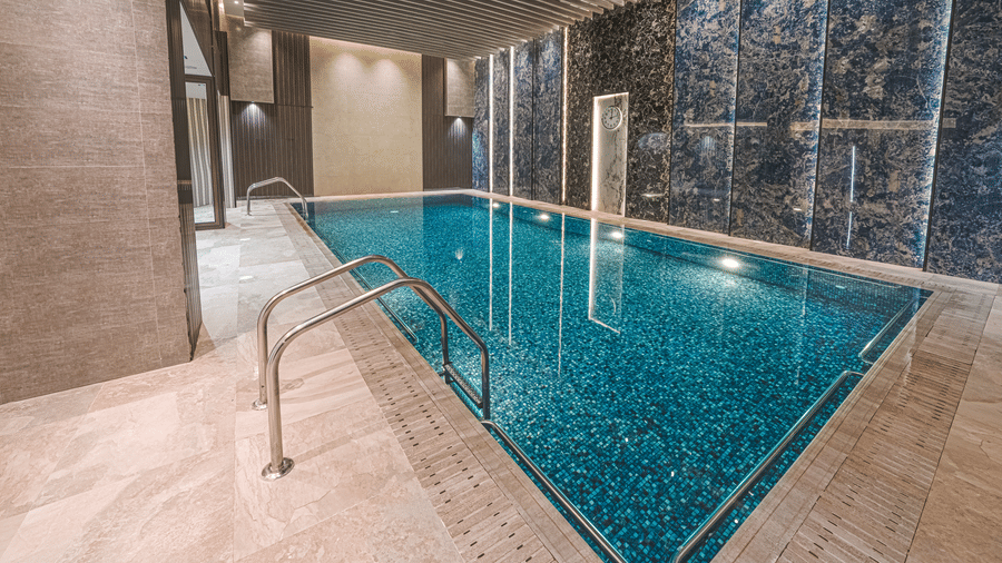 Wide view of the indoor swimming pool at Grand Iva Hotel, Abha, seen with a clean and spacious deck, pool steps, a panelled wall behind it with a clock on it.