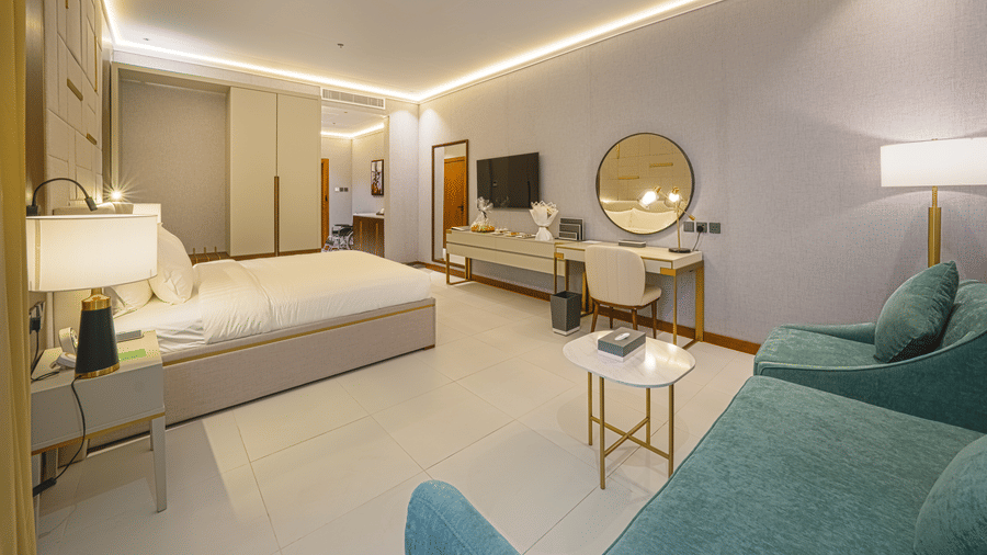 Deluxe King ADA-friendly bedroom corner view showcasing the plush bed facing the wall-mounted TV, the work desk situated beside an upholstered couch in a corner | Grand Iva Hotel, Abha