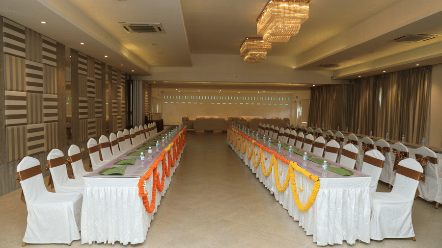A long indoor banquet hall with multiple long tables arranged for an event, featuring white chairs with sashes and colourful table runners - Grande Bay Resort & Spa, Mamallapuram