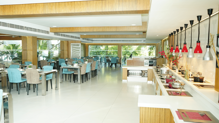 Wide-angle view of L’attitude 49 restaurant in Mahabalipuram featuring blue chairs, large windows, buffet counters, hanging lamps, and bright interiors - Grande Bay Resort & Spa, Mamallapuram
