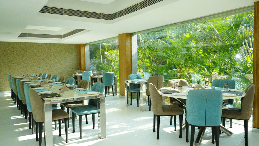 Indoor dining area with elegant table arrangements and large windows showcasing green coconut trees - Grande Bay Resort & Spa, Mamallapuram