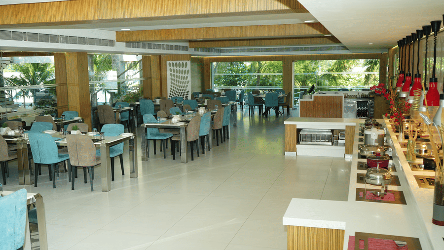 Wide-angle view of L’attitude 49 restaurant in Mahabalipuram featuring blue chairs, large windows, buffet counters, hanging lamps, and bright interiors - Grande Bay Resort & Spa, Mamallapuram