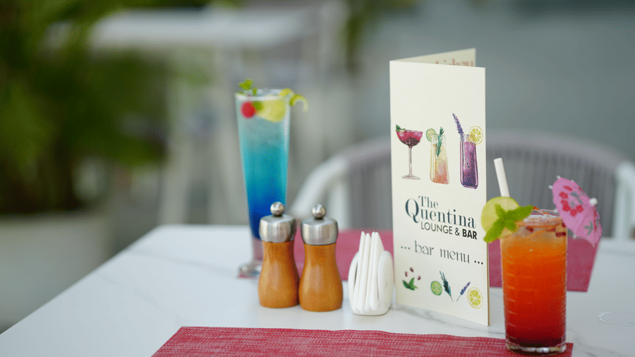 Outdoor drink table setup from The Quentina Bar featuring colourful cocktails, menu card, and condiments - Grande Bay Resort & Spa, Mamallapuram