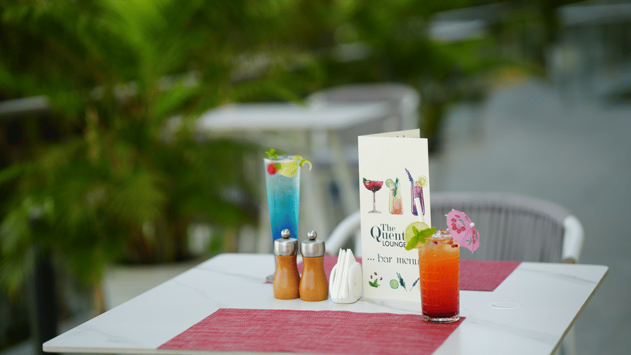 Outdoor dining table with a red drink, a blue drink, menu card and condiments at Grande Bay Resort & Spa, Mamallapuram.