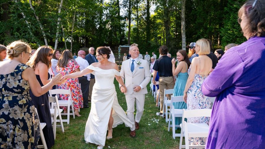 At Hartstone Inn, a newlywed couple walks down the aisle holding hands, surrounded by standing guests who are smiling, clapping, and reaching out to greet them.