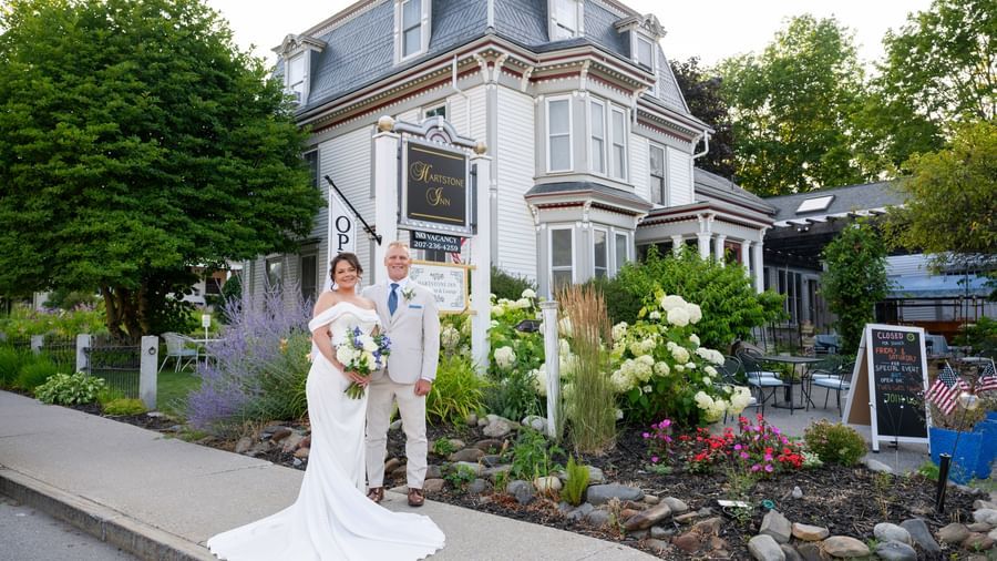 A bride in a dress and a groom in a suit stand in front of the Hartstone Inn sign, with flowers, a tree, and the building visible in the background.