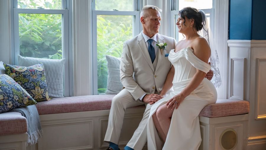 Bride and groom sitting on a window seat at Hartstone Inn, holding hands and looking at each other, with pillows and greenery visible through the windows.