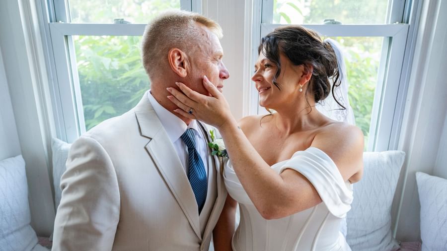 A man in a suit and a woman in a wedding dress are sitting by a window, facing each other, with the woman touching the man's face at Hartstone Inn.