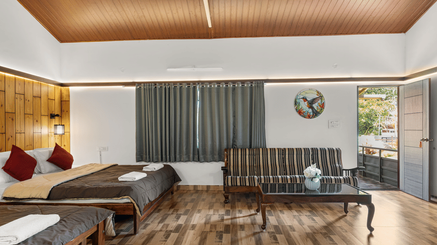 Bedroom with a bed dressed in white linen and red cushions, a grey sofa, a wall-mounted television and a wooden panelled ceiling overhead.