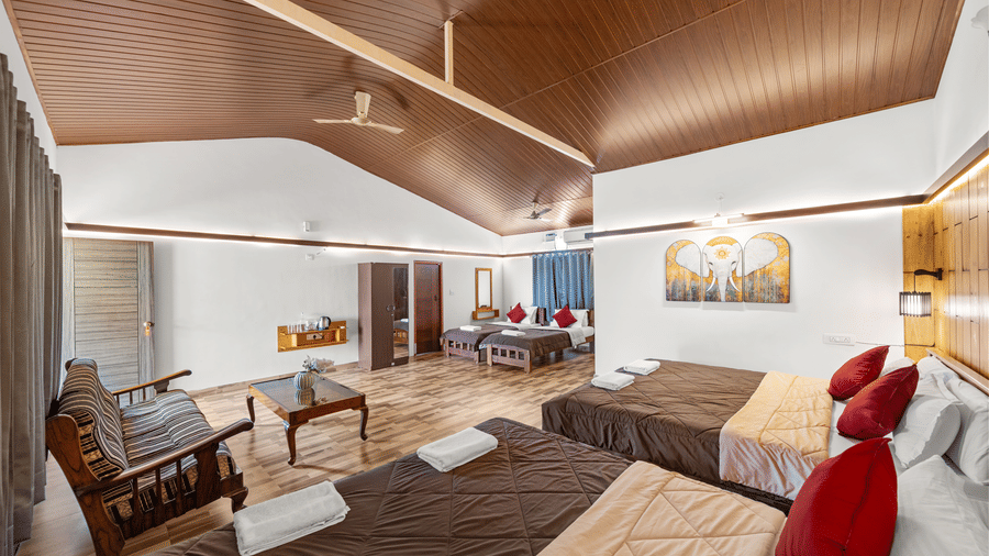 Open living area with beds dressed in brown linen and red cushions, wooden furniture, a wooden beamed ceiling and warm ambient lighting nearby.