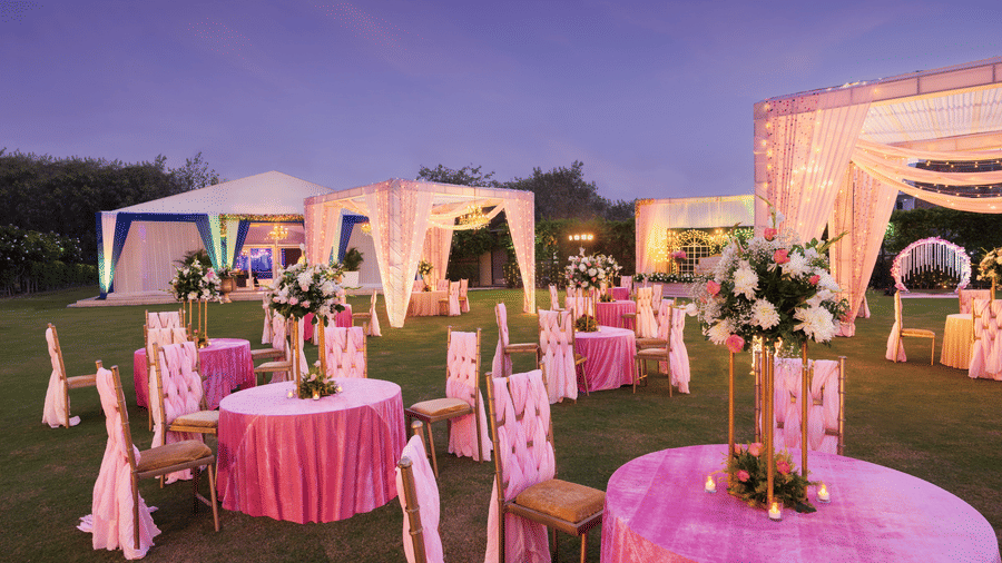 Clusters of chairs and tables with pink covers and canopies lit up with decorative lights during the evening - The Hideaway Banquets & Lawns, Greater Noida
