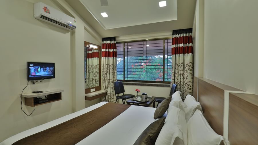 Extended view of the queen size bed facing the wall-mounted TV in the Deluxe Room of Highland Resort, that also features drawn curtains offering views of the trees outside.