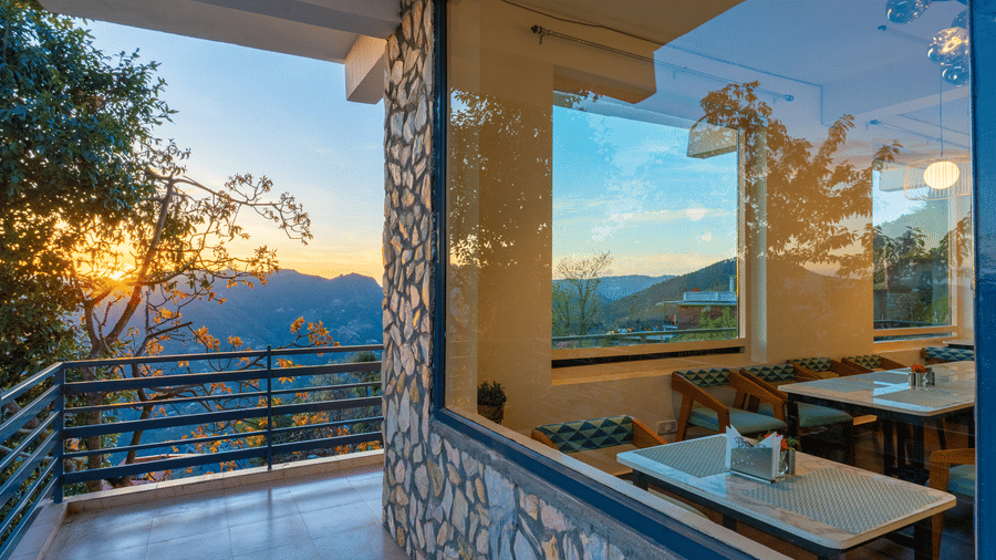 A view through a large glass window of an indoor dining area overlooking a valley at Himalayas Resort By The Lake Hill, Mukteshwar.