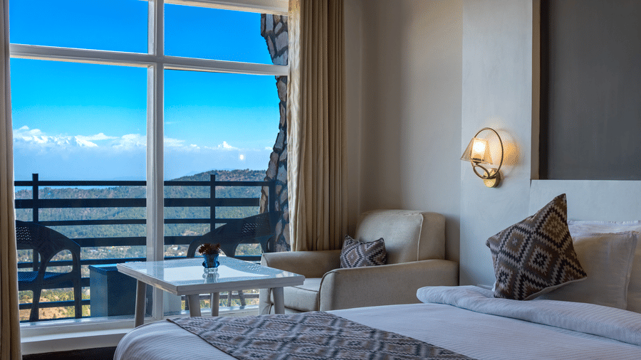 View of a hotel room window with a blue sky outside, featuring an armchair and a side table at Himalayas Resort By The Lake Hill, Mukteshwar.