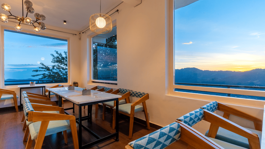 A dining table in a cafe with large windows offering a view of the sun setting over the mountains at Himalayas Resort By The Lake Hill, Mukteshwar.