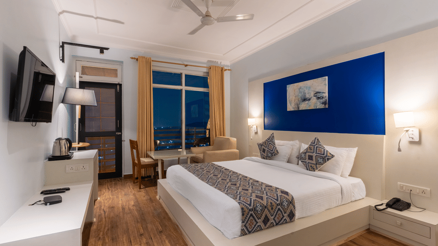 Modern hotel bedroom featuring a double bed, blue accent wall, wall-mounted TV, and wooden flooring at Himalayas Resort By The Lake Hill, Mukteshwar.
