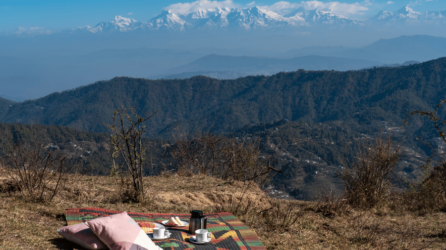 A picnic blanket and cushions spread on a grassy ridge overlooking the snow-capped Himalayan peaks at Himalayas Resort By The Lake Hill, Mukteshwar.
