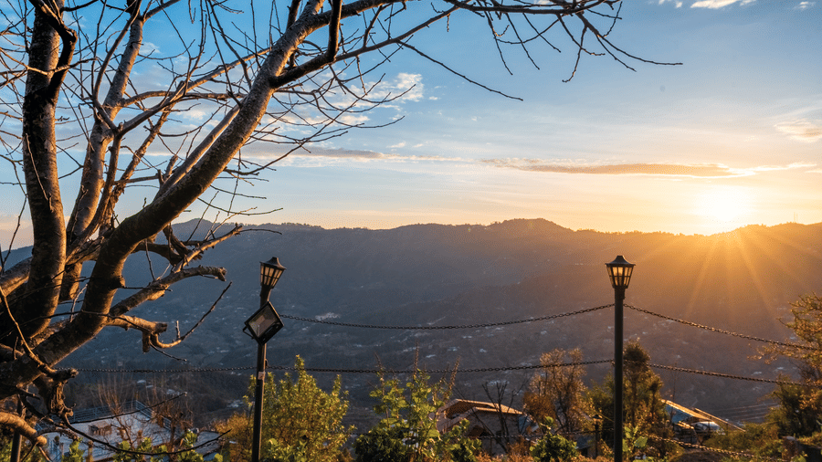A scenic view of the sun setting over distant mountain ridges seen through silhouetted tree branches at Himalayas Resort By The Lake Hill, Mukteshwar.
