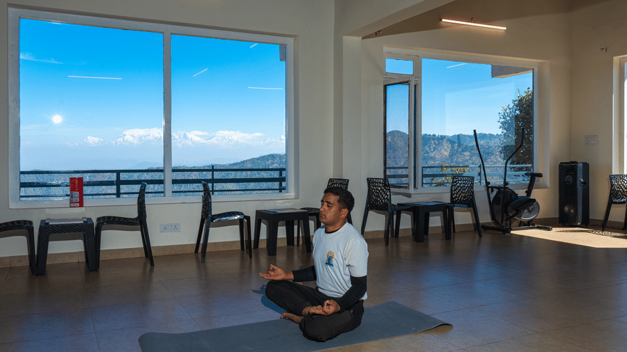A guest practicing yoga on a mat in a bright room with large windows showing a mountain view at Himalayas Resort By The Lake Hill, Mukteshwar.