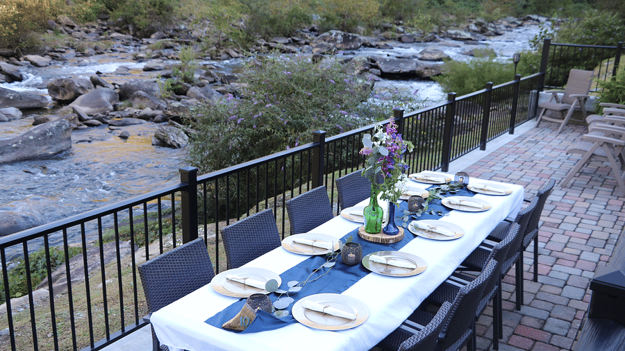 a long dining table decorated with a flower vase along with well arranged cutlery and glassware overlooking the serene river views from the railings - Historic Tapoco Lodge
