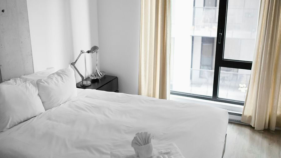 A hotel room with white sheets, side table and lamp next to the window