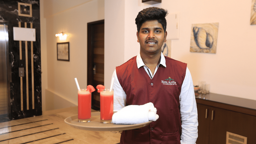 A man holding a tray that has two glasses of drink on it at Hotel Altitude, Madikeri.