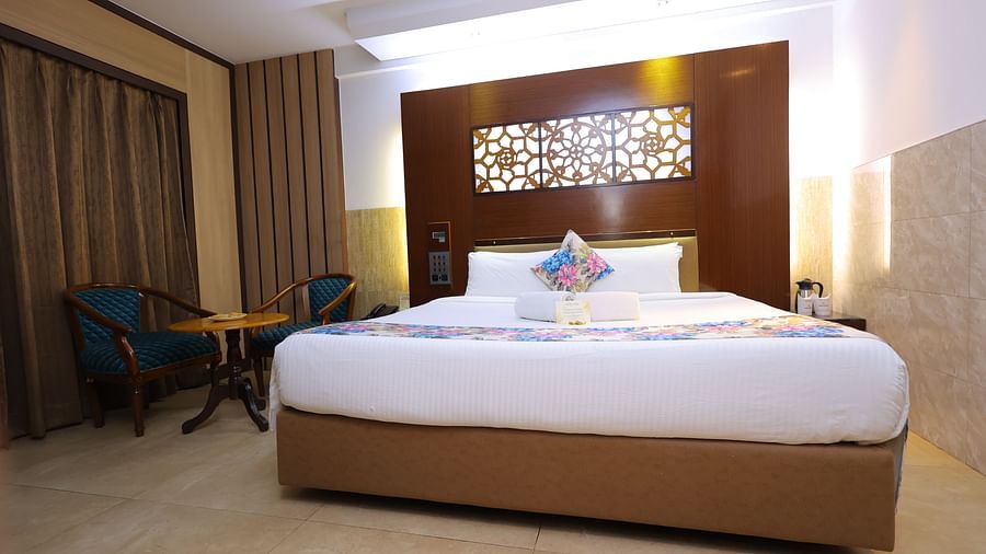 Lower angle view image of a deluxe room at Hotel Amar with a bed, table, chair and huge window with curtains. 