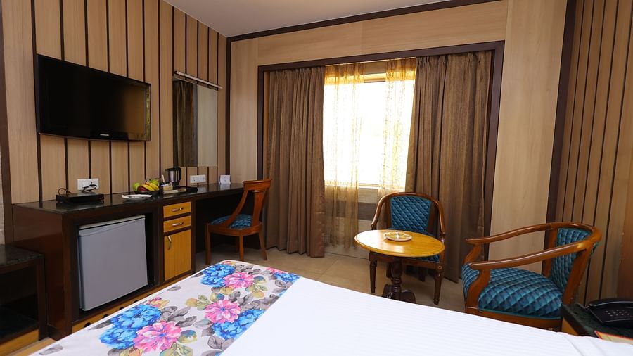 Side view image of a deluxe room at Hotel Amar with a bed, table, TV and huge window with curtains. 