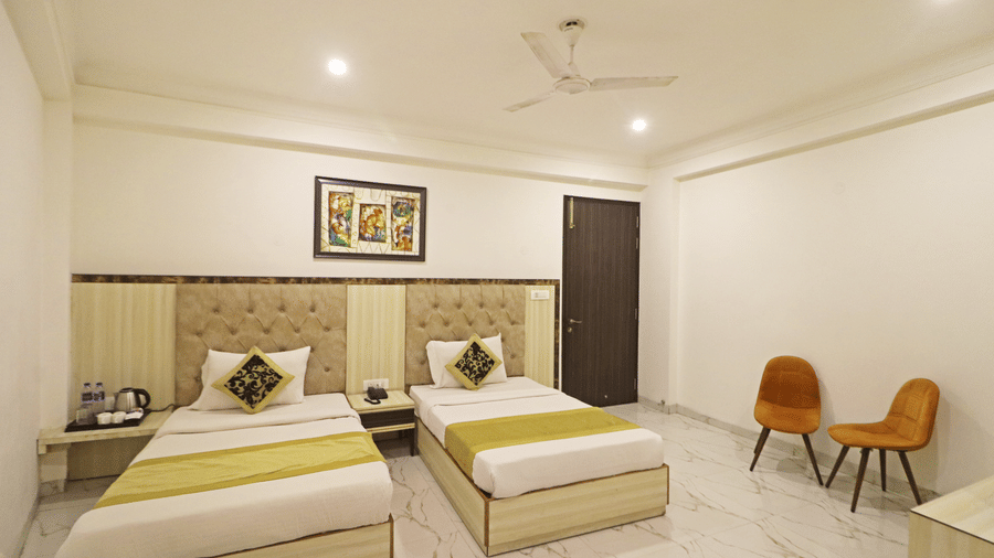 A twin guest room at Hotel Apple Park Delhi with two single beds featuring lime green runners, a central headboard, and two orange chairs against a white wall.