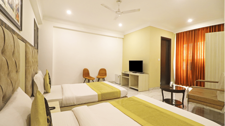 A side-angle view of a twin room at Hotel Apple Park Delhi showing the beds, a television on a light wood stand, and bright orange curtains.