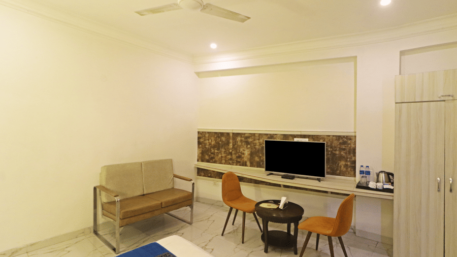 A guest room at Hotel Apple Park Delhi showing a long work desk with a television, two orange chairs, and a metal-framed sofa.