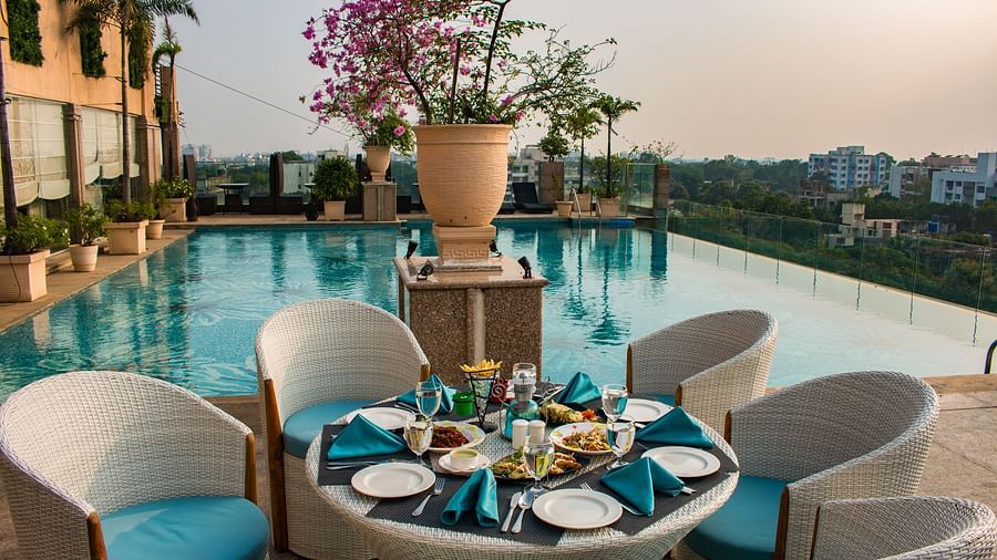 An outdoor dining at Hotel Ashok Nagpur featuring a round table with place settings for four, surrounded by cushioned chairs, and a swimming pool and cityscape in the background. 