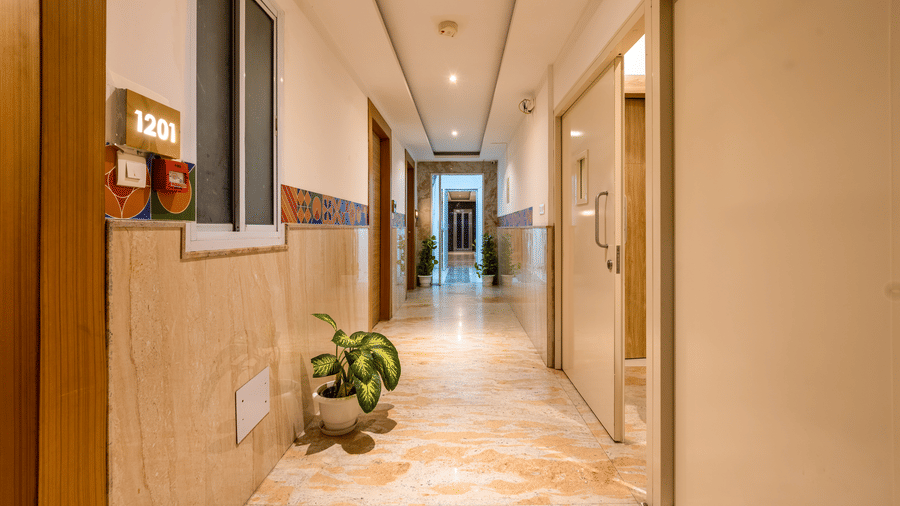 A quiet corridor at Hotel Corporate Suites with marble-like tiled flooring leading towards a lift, decorated with potted plants against plain walls