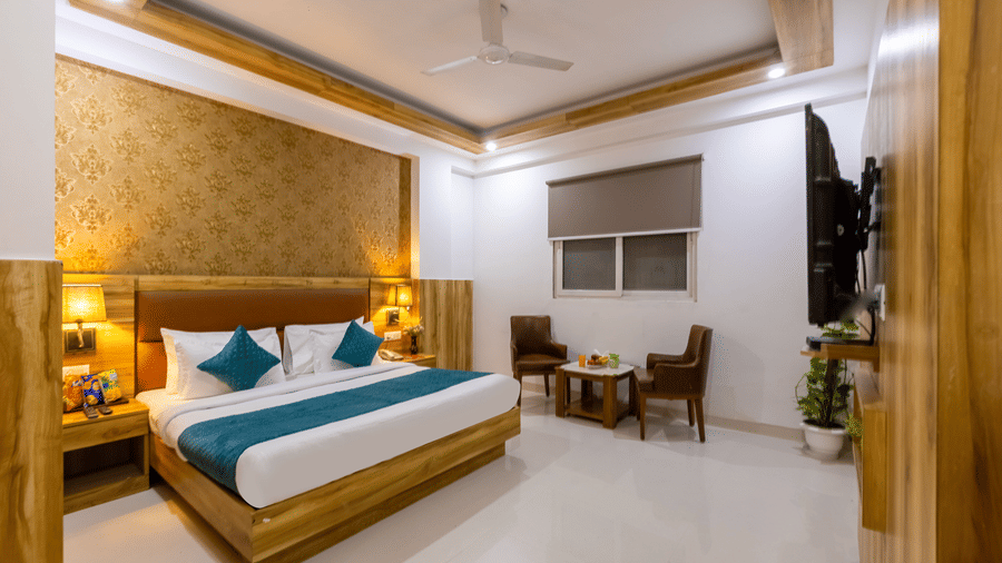 A well-lit Deluxe Room showcasing a double bed against a patterned wall facing the TV and a seating space by the window