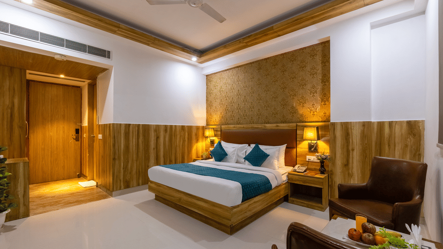 A bright Deluxe Room featuring a plush bed with teal accents against patterned wallpaper, a cosy brown leather armchair, and a fruit basket on a small table at Hotel Corporate Suites