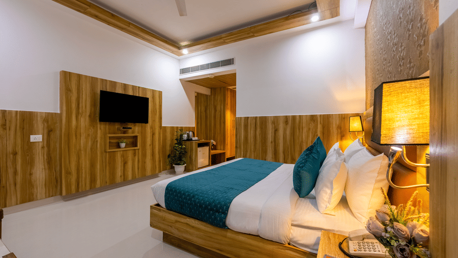 Deluxe Room showcasing a bed with a teal runner and matching accent cushions facing a flat-screen TV and a minibar unit, and soft lighting at Hotel Corporate Suites