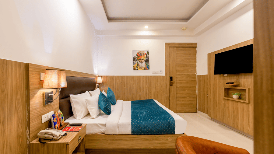 Deluxe Room at Hotel Corporate Suites featuring a plush bed facing the TV mounted on the wall and a nightstand with a telephone on it with a lamp lit over it