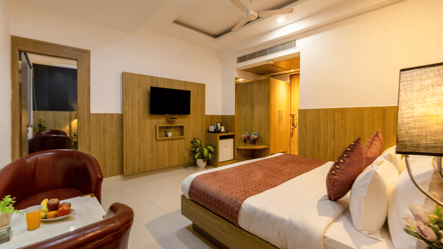 Executive Room at Hotel Corporate Suites under warm lighting featuring a bed facing the TV on the wall with minibar below it and a seating space with 2 chairs and fruits kept on the coffee table