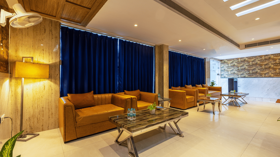 Lobby seating area furnished with comfortable tan couches, a wooden table holding bottled water, and floor-to-ceiling blue curtains at Hotel Corporate Suites